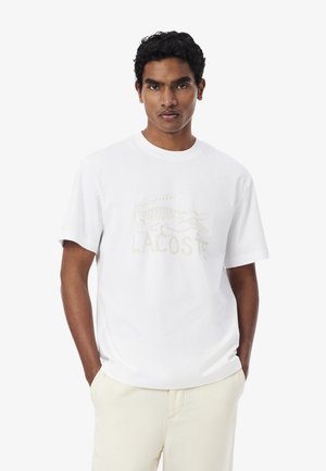 Hombre con cabello rizado vistiendo una camiseta blanca de Lacoste y pantalones beige claro, de pie con las manos en los bolsillos contra un fondo liso.