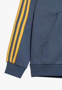Blue sweatshirt sleeve with three vertical yellow stripes and a front pocket on a white background.