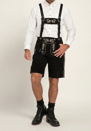 Zwarte suède lederhosen met decoratieve borduurwerk, verstelbare schouderbanden en metalen beslag. Gecombineerd met een wit overhemd met knopen.