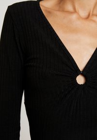 Svart ribbat topp med en djup V-ringning och samlingsdetalj runt en central cirkulär utskärning. Har långa ärmar och texturerad tyg.