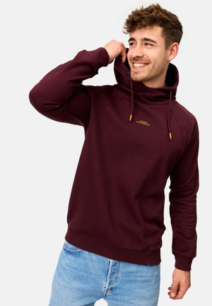 Bordeaux hoodie met trekkoord, voorzien van ribbels aan de manchetten en onderzoom. Tekst "ICONIC EXPERIENCE" in het geel op de borst, gecombineerd met lichtblauwe spijkerbroek.