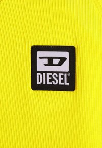 Jasnożółty ribbed materiał z czarną prostokątną łatą, na której znajduje się napis "DIESEL" białymi literami oraz stylizowane logo w rogu.