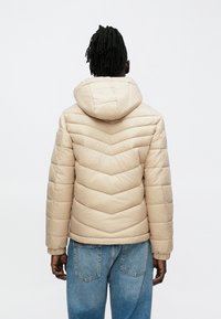 Μπεζ μονω μένο μπουφάν πάπλωμα με σχέδιο ζιγκ-ζαγκ, ενσωματωμένη κουκούλα και ελαστικές μανσέτες. Συνδυασμένο με ανοιχτό μπλε τζιν παντελόνι.