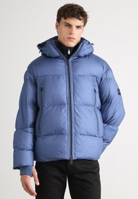 Giacca invernale imbottita di un blu chiaro opaco, con cappuccio, chiusura a zip e due tasche laterali con zip. Texture morbida con cuciture orizzontali.