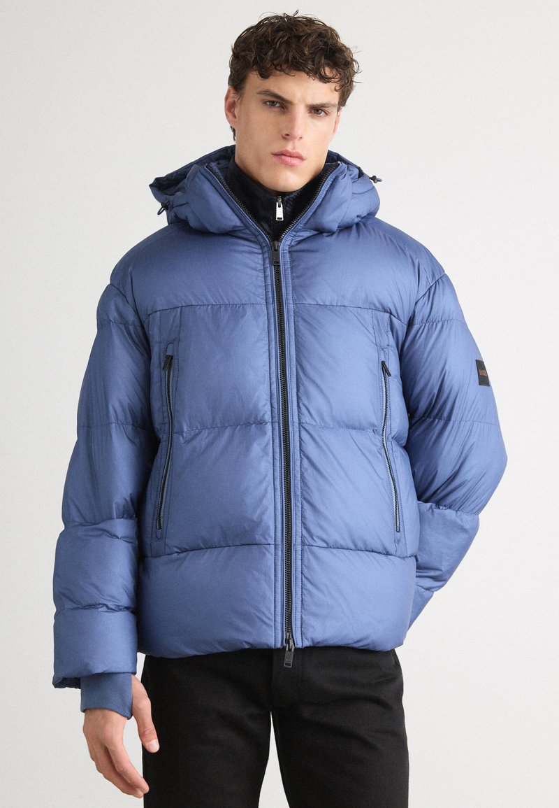 Giacca invernale imbottita di un blu chiaro opaco, con cappuccio, chiusura a zip e due tasche laterali con zip. Texture morbida con cuciture orizzontali.