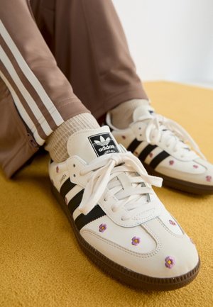 Baskets blanches Adidas à rayures noires avec une petite broderie de fleur violette, portées avec des chaussettes beiges et un pantalon de survêtement marron, sur une surface jaune.
