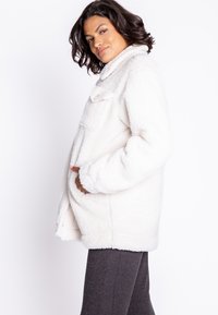 Hvid fleecejakke med høj krave og frontlomme, lavet af et blødt, tekstureret stof og med en løs pasform, iført over mørke leggings.