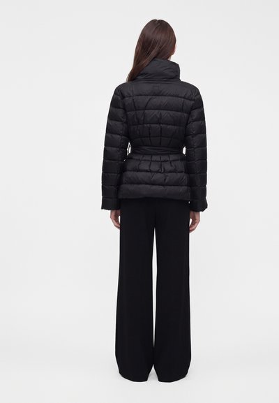 Femme portant une doudoune noire ceinturée et un pantalon noir ample, debout, dos tourné, sur un fond clair uni.