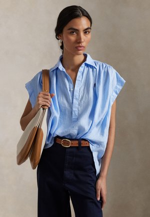 Γυναίκα που φοράει φαρδιά ανοιχτομπλε κοντομάνικη μπλούζα, σκούρο παντελόνι, καφέ ζώνη, κρατώντας τσάντα ώμου σε μπεζ και καφέ χρώμα σε απλό φόντο.