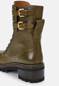 Stivaletto alla caviglia in pelle verde oliva con fibbie color oro, suola robusta e tacco impilato. Presenta un marchio in rilievo sul lato.