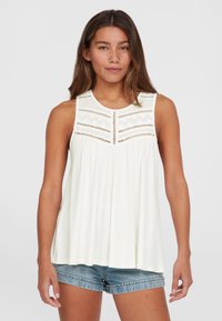 Top bianco senza maniche con colletto rotondo, caratterizzato da ritagli decorativi e dettagli ricamati sul petto, abbinato a pantaloni corti di denim.