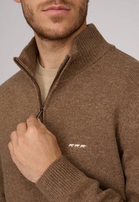 Un pull zippé marron avec un col haut, fabriqué dans un matériau texturé. Présente une petite broderie d'animal blanc sur la poitrine.