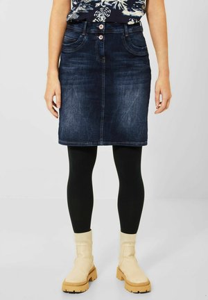 Vrouw die een donkerblauwe spijkerrok, zwarte legging en crèmekleurige enkellaarsjes draagt, staand tegen een effen lichte achtergrond.