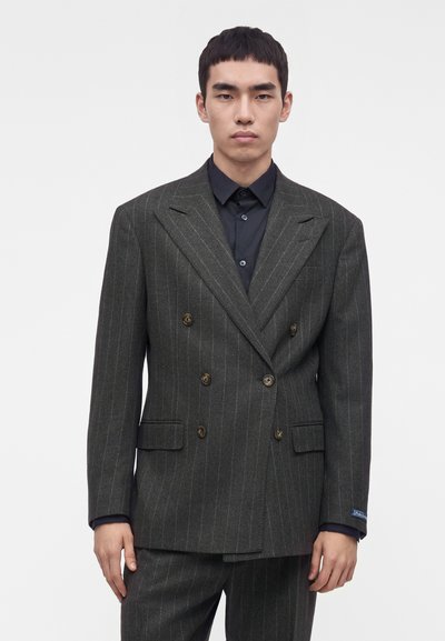 Jeune homme portant une veste de costume croisée à rayures gris foncé sur une chemise noire boutonnée, debout devant un fond blanc uni.