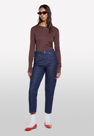 Chemise marron à manches longues, jeans en denim foncé taille haute, ballerines rouges, chaussettes blanches mi-mollet et lunettes de soleil marron. Textures lisses avec une coupe décontractée.