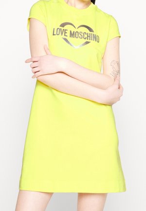 Kvinde iført en knalddysergul kortærmet kjole med "LOVE MOSCHINO" og hjerte-logo, med armene krydset, tatovering synlig på højre underarm.