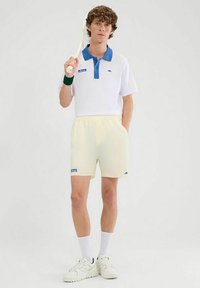 Weißes Polo-Shirt mit blauem Kragen, gelben Shorts, grünem Handgelenkband und weißen Turnschuhen, mit einem Tennisschläger in der Hand. Einfaches Design.