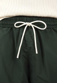 Cordon blanc noué en nœud sur un pantalon taille élastique vert foncé, associé à un pull blanc côtelé rentré.