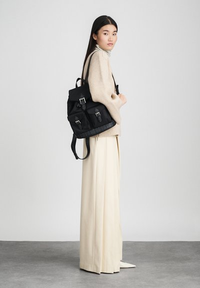 Mujer con cabello largo y oscuro, vestida con un suéter beige, pantalones amplios crema, zapatos blancos, llevando una mochila de cuero negra, de pie de lado sobre un suelo gris.