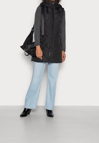 Schwarz gesteppte Weste mit Kapuze, mit einem Reißverschluss vorne und Chevron-Design, getragen über einem grauen Pullover und kombiniert mit hellblauen Schlagjeans und schwarzen Stiefeln.