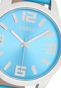 Montre avec un cadran bleu et finition brossée, index des heures argentés et boîtier rond. Dispose d'un logo de marque argenté et d'un bracelet bleu assorti.