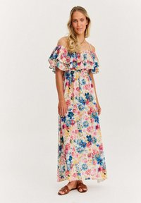Kwiatowa sukienka maxi z odkrytymi ramionami i falbanami, ozdobiona niebieskimi, różowymi i żółtymi kwiatami na jasnym tle. Noszona z brązowymi sandałami.