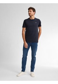 Camiseta de manga corta en color marino, hecha de algodón. Presenta un cuello redondo y un pequeño logo en el pecho. Combinada con jeans azules y zapatillas blancas.