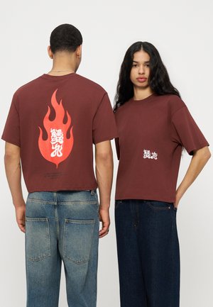 Δύο άτομα φορούν καφέ υπερμεγέθη T-shirts με λευκούς ασιατικούς χαρακτήρες· το ένα δείχνει ένα μεγάλο κόκκινο σχέδιο φλόγας στην πλάτη, το άλλο στέκεται με το πρόσωπο προς τα εμπρός.