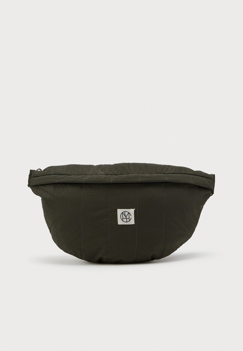 Σκούρο πράσινο επενδεδυμένο ύφασμα μέσης τσάντα με ένα μικρό λευκό λογότυπο κεντραρισμένο στο εμπρός μέρος σε έναν απλό φόντο.