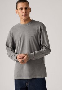 Šedé tričko s dlhým rukávom vyrobené z bavlny, s okrúhlym výstrihom, viditeľnou potlačou loga na hrudi a rebrovanými manžetami.