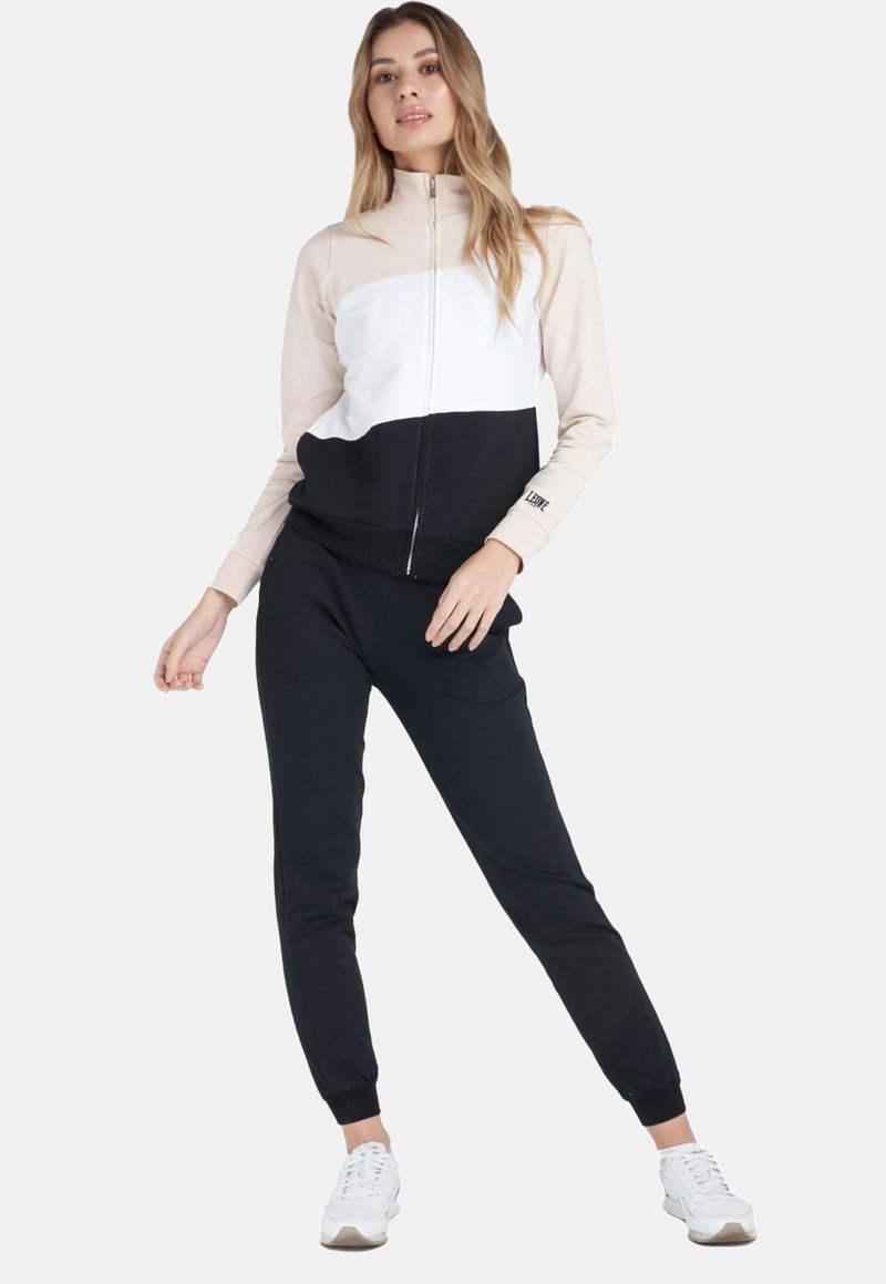 Une femme porte une veste zippée à blocs de couleurs, un pantalon de jogging noir et des baskets blanches sur un fond blanc uni.
