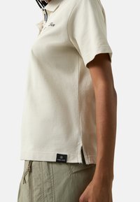 Polo crème à manches courtes, doté d'un col et d'ouvertures latérales. Logo brodé en haut à gauche. Porté avec un pantalon vert olive.