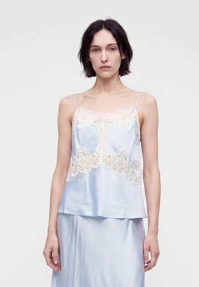 Femme aux cheveux courts et foncés, portant un caraco en satin bleu clair avec des détails en dentelle beige et une jupe assortie sur un fond uni.