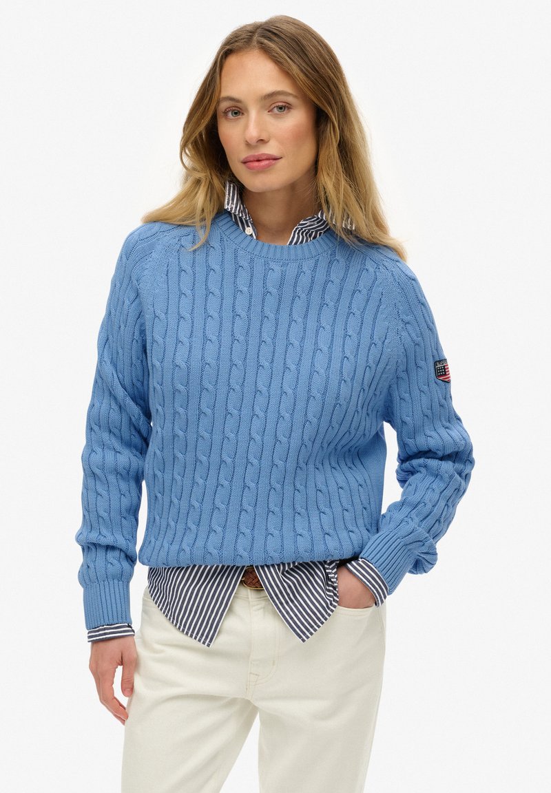 Pull en maille torsadée bleu clair avec un col rond. Il présente des poignets et un ourlet côtelés, superposé à une chemise à col rayée bleu marine et blanche.