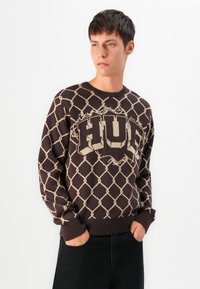 Young man with short dark hair wearing a dark brown sweater with beige chain-link pattern and large block letters across the chest.
