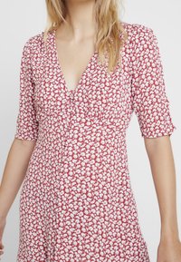 Rode bloemenjurk met korte mouwen, v-hals en geplooide detail bij de buste; witte bloemenpatronen op een textuurstof.