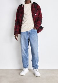 Rood en zwart geruite shirt met een witte sherpa kraag, gecombineerd met een beige T-shirt en lichtblauwe tapered jeans. Witte sneakers maken de look af.