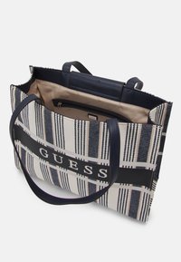 Tygväska med marinblå och krämfärgad rutigt mönster, dubbla marinblå handtag och ett svart band med varumärkesnamnet "GUESS". Insida med dragkedjeficka.