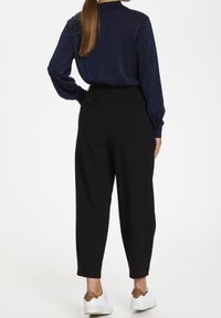 Navy glinsterend shirt met lange mouwen en hoge hals, gecombineerd met zwarte high-waisted, taps toelopende broeken en witte sneakers met bruine accenten.