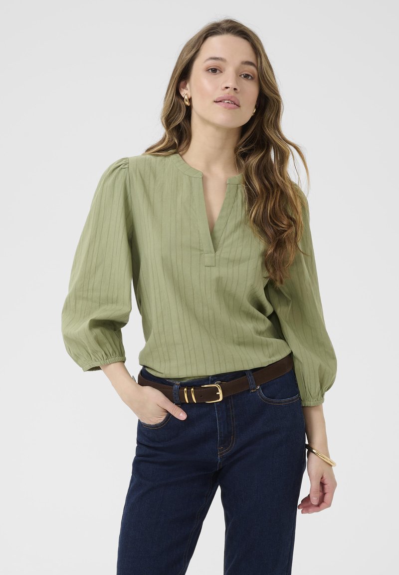 Blusa verde pallido con strisce verticali, maniche a tre quarti plissettate e scollatura a V, abbinata a jeans blu scuro e una cintura marrone.