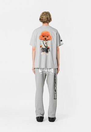 Camiseta gris con un gráfico de un perro de dibujos animados en un traje de artes marciales. Combinada con pantalones de chándal grises que muestran un logotipo. Diseño casual.