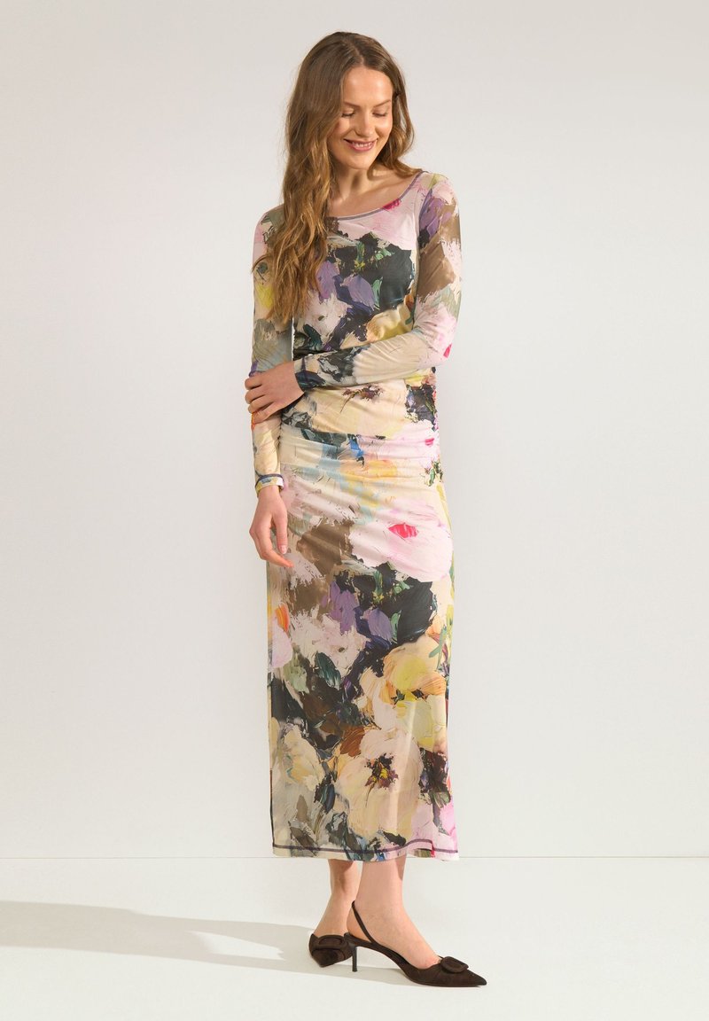 Femme portant une longue robe fleurie multicolore à manches longues, debout les bras croisés et chaussant des talons slingback noirs pointus.