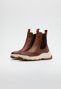 Botas de tobillo de cuero marrón con paneles elásticos, detalles perforados y una suela gruesa blanca crema. Tiradores rojos en la parte superior.