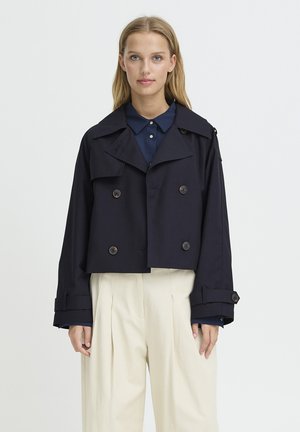 Giovane donna che indossa una giacca doppiopetto corta blu navy su una camicia scura con colletto e pantaloni plissettati crema a vita alta, in piedi.