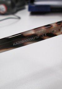 Bruine schildpad kunststof zonnebril met een slank, gebogen ontwerp. Gegraveerde tekst op de poot: "KA00B0000A21D" en "CE"-merk zichtbaar.