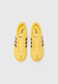 Geltoni sportbačiai su rudomis juostelėmis, apvaliu nagu ir raišteliais. Pagaminti iš tekstilės, turinčios lygų paviršių ir subtilų ženklinimą viduje.