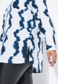 Biała koszula z niebieskim wzorem tie-dye, długie rękawy, kieszeń na piersi, rozcięcia po bokach, zestawiona z czarnymi skórzanymi leggingsami. Gładka tekstura materiału.