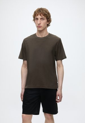 Ung mand med krøllet lysebrunt hår iført en mørkebrun kortærmet skjorte og sorte shorts, står mod en ensfarvet hvid baggrund.