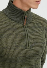 Grüner Zip-Hoody mit hohem Kragen, strukturiertem Strickmuster und einem braunen Lederriegel mit dem Markennamen nahe dem Reißverschluss.