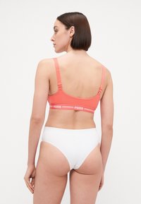 Coral sports bra with adjustable straps, featuring a white branded band. Paired with white brief, showing a smooth, minimalist design.